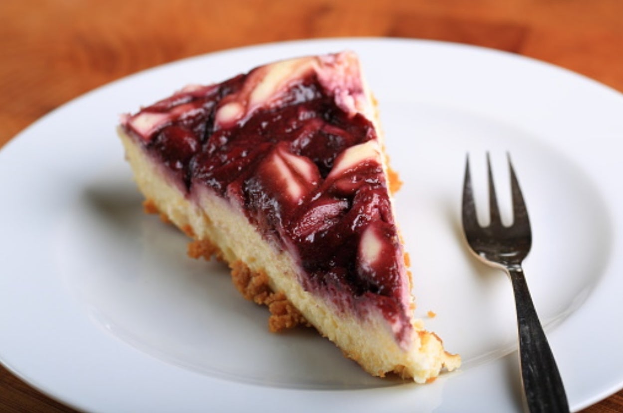 A slice of cheesecake topped with swirled berry compote on a white plate, accompanied by a fork