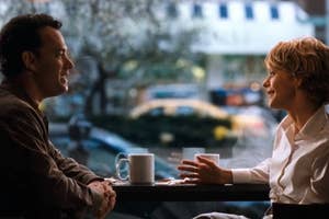 Two people sit at a café table, engaged in conversation, with coffee cups in front of them and a blurred outdoor view in the background