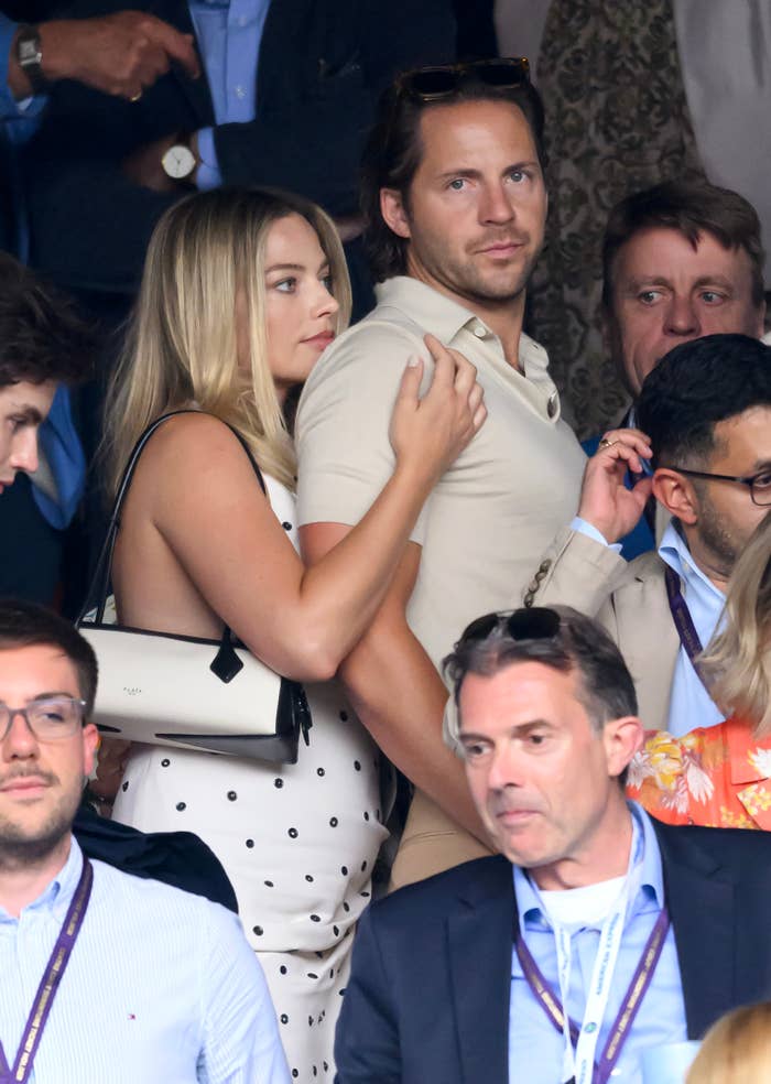 A personage mates is successful a assemblage astatine an outdoor event. The pistillate wears a polka dot formal and holds a achromatic bag, portion the antheral is successful a casual beige outfit