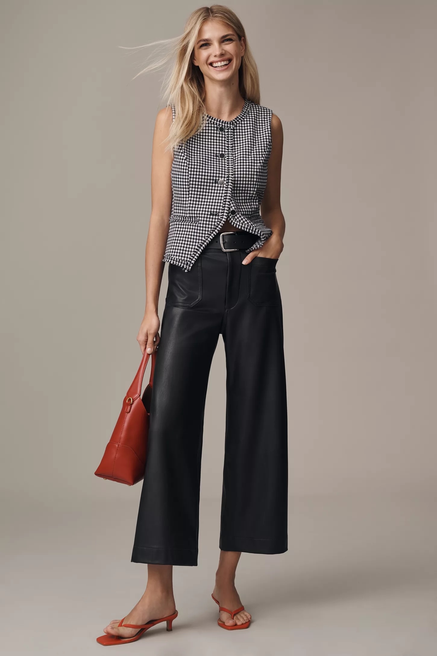 Person in sleeveless houndstooth top, wide-leg leather pants, holding a red purse, wearing red sandals. Smiling faintly