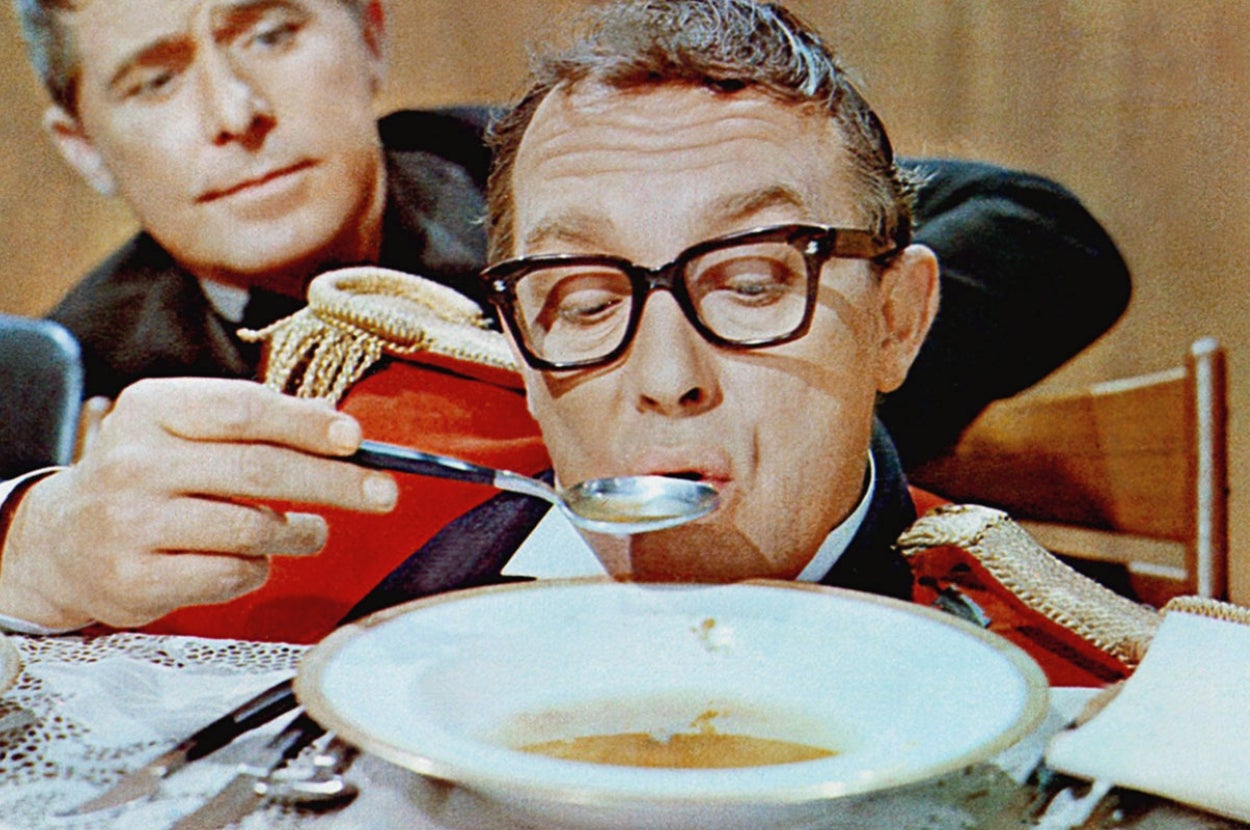 Eric Morecambe in "The Intelligence Man" with glasses sipping soup from a spoon at a table, with another man in the background watching intently
