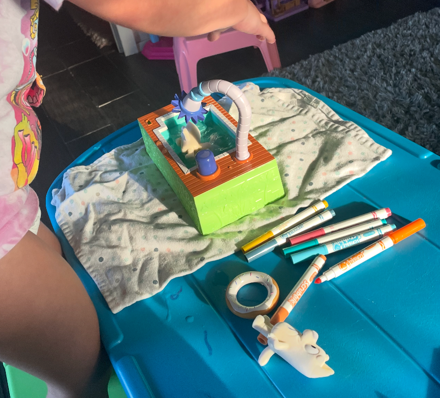 Child playing with a artifact  washing instrumentality   connected  a table, surrounded by markers and a rotation  of tape