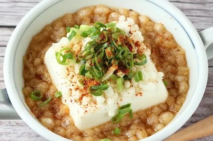 茶碗に入った豆腐料理。豆腐の上に葱と香辛料がトッピングされ、スープに浮いている。木製のスプーンが横に置かれている。