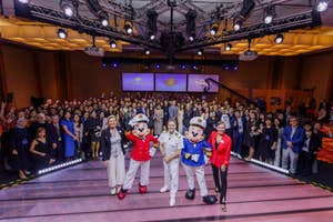 Group photo of three costumed characters and four people in formal attire posing with a large crowd at a themed event