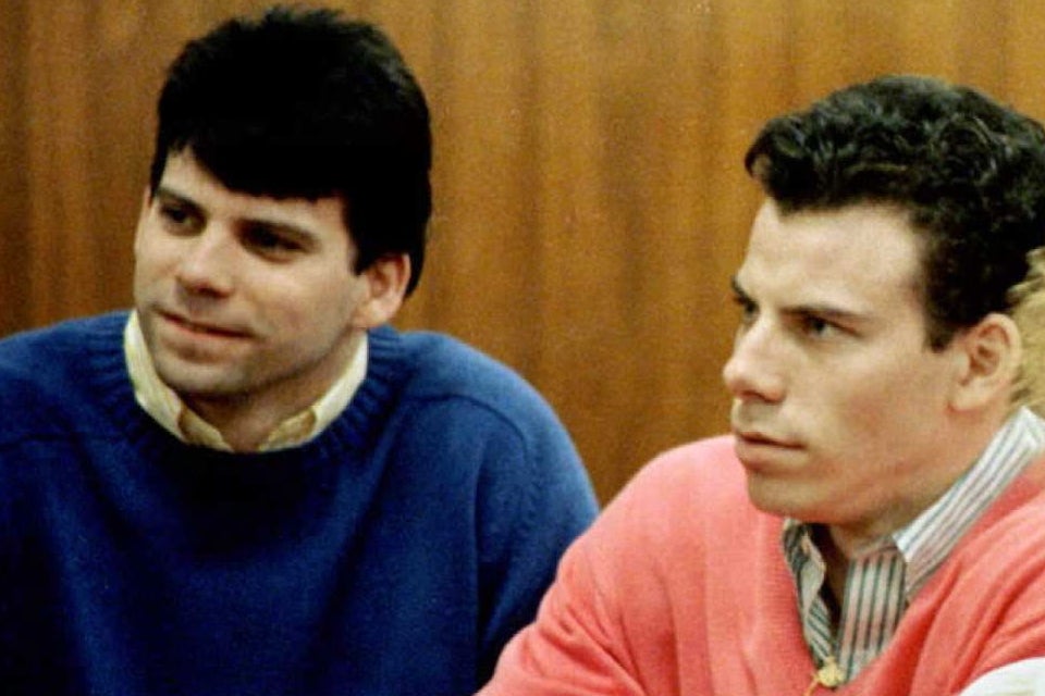 Lyle and Erik Menendez in court, wearing casual sweaters, appear attentive and serious, seated beside a woman with curly hair