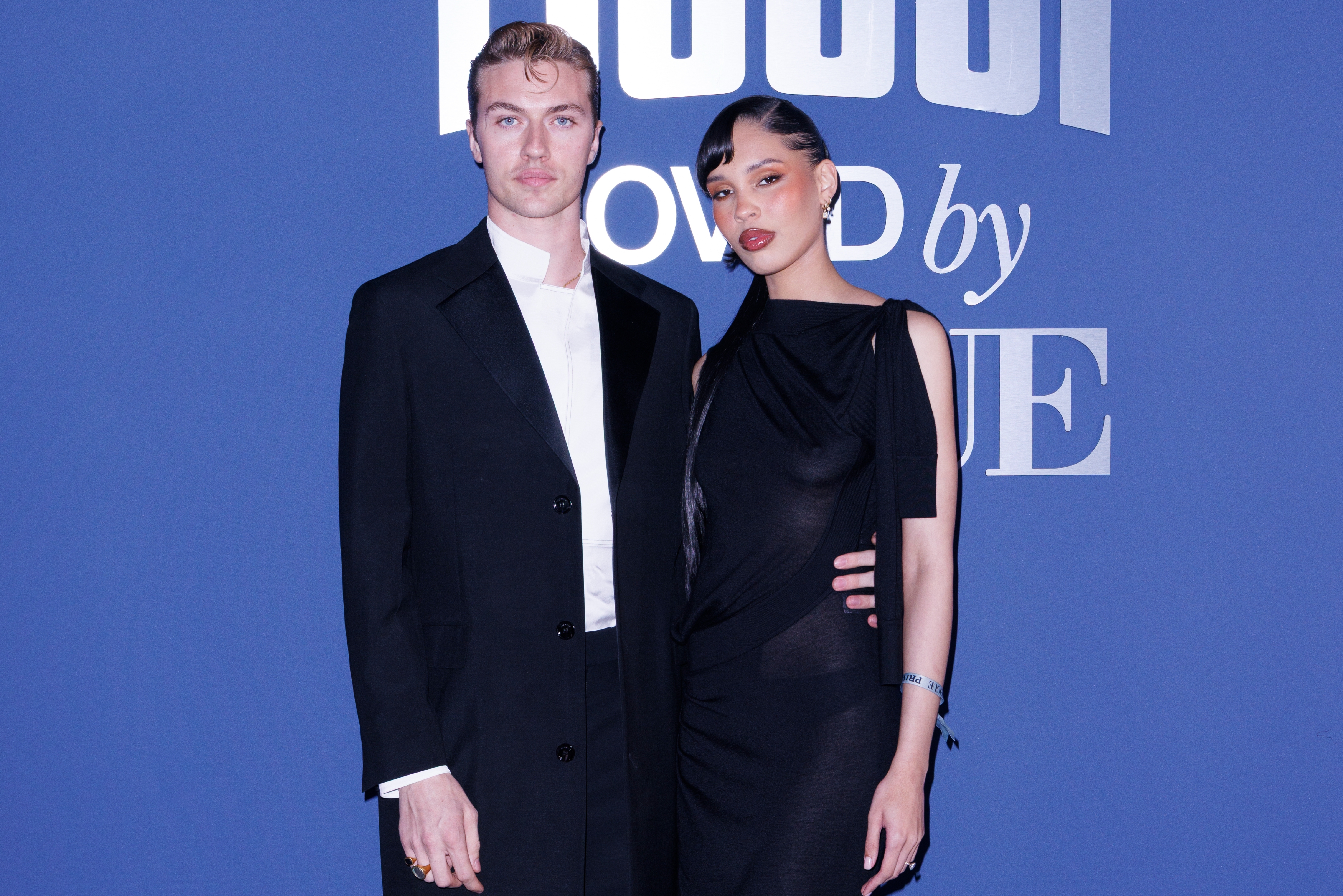 Lucky Blue Smith and Nara Smith posing together at an event, dressed in stylish formal attire. A logo is visible in the background
