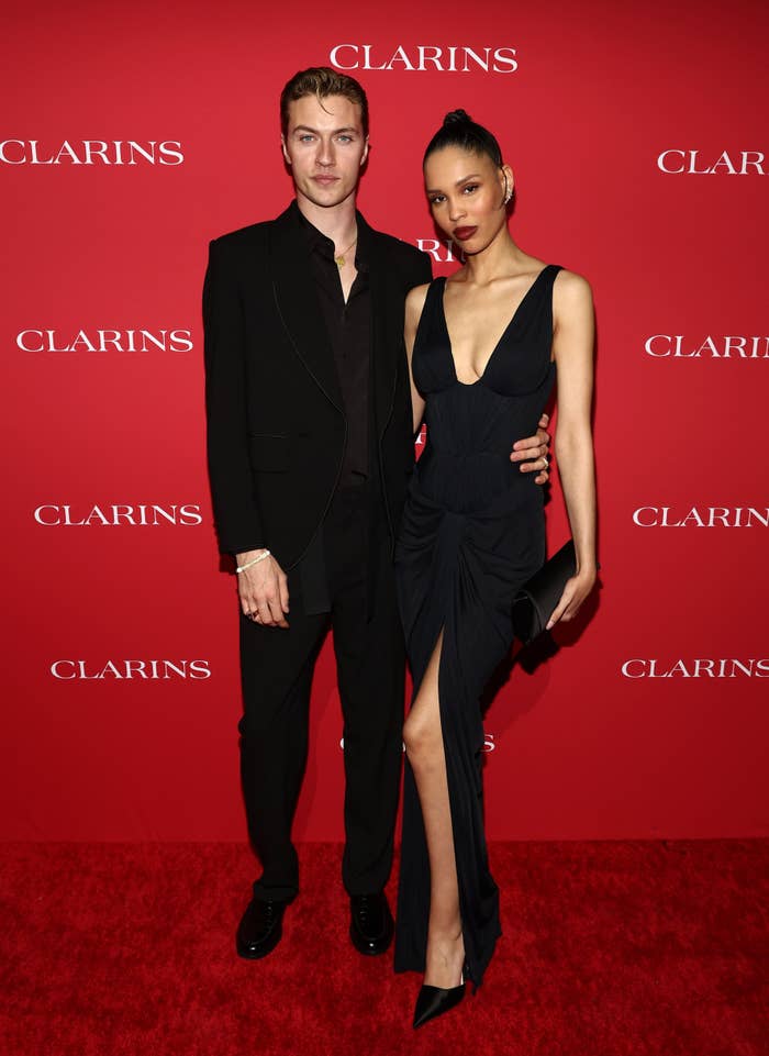 Lucky Blue Smith and Nara Smith posing connected a reddish carpet. One is wearing a acheronian suit, the different a sleeveless gown with a slit. Red backdrop with "Clarins" logos