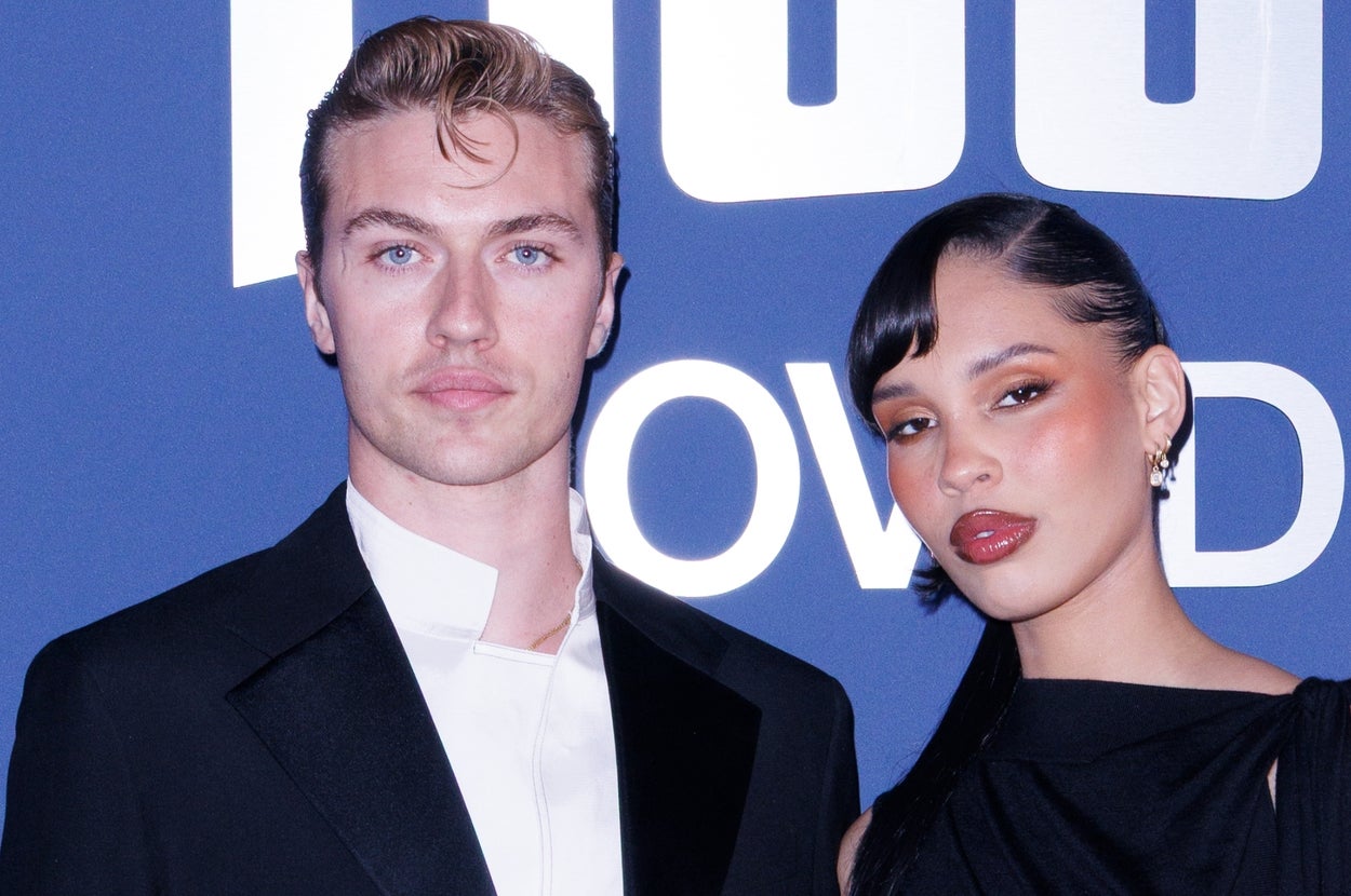Two individuals on a red carpet; one wears a classic black suit, and the other sports a sleek, form-fitting black dress with a draped design