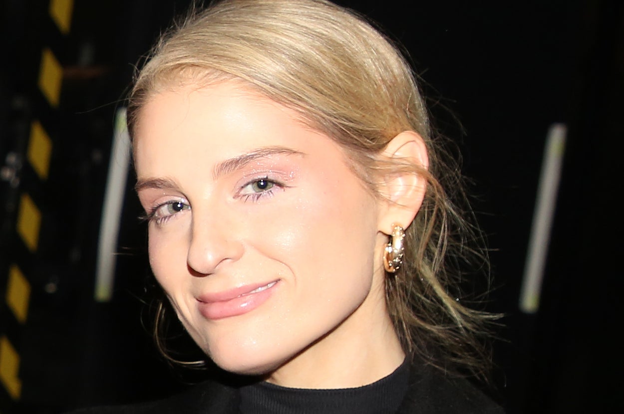 A person in a black coat with gold buttons, smiling slightly at the camera. They have accessorized with gold hoop earrings