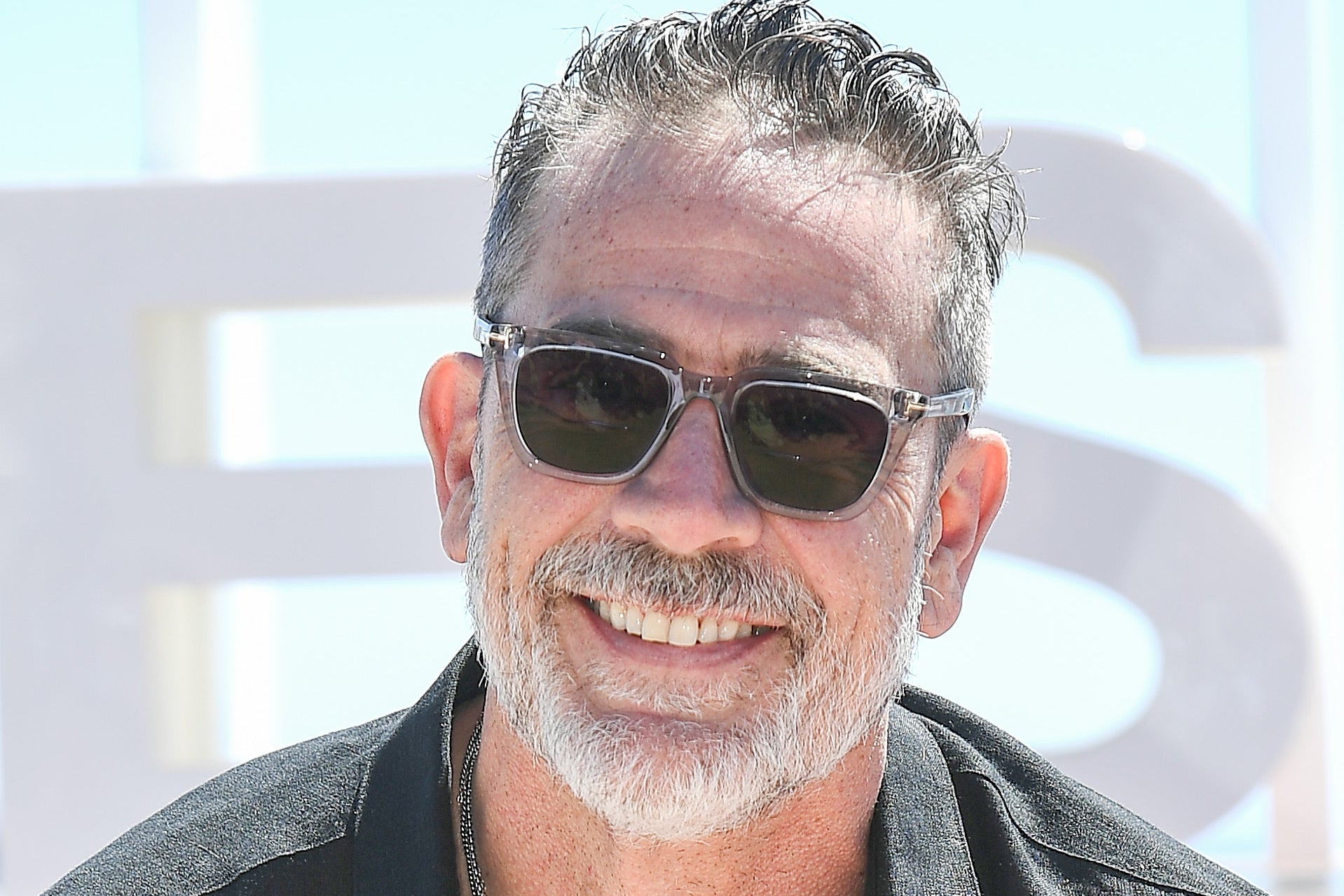 Man in a casual dark shirt, sunglasses, smiling outdoors. Short hair, beard, and necklaces visible