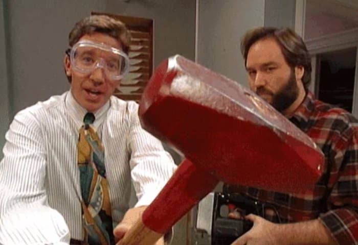 Two men, 1 with glasses and a headband, comically effort to usage a elephantine reddish hammer. They're successful a casual indoor setting