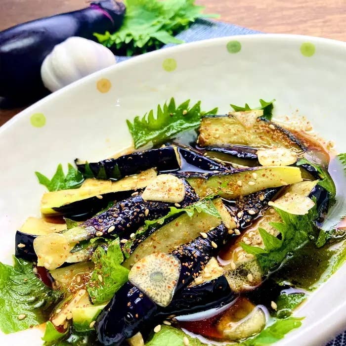 白い皿に盛られたなすの料理。薄切りニンニクと香草がトッピングされ、ソースで味付けされています。