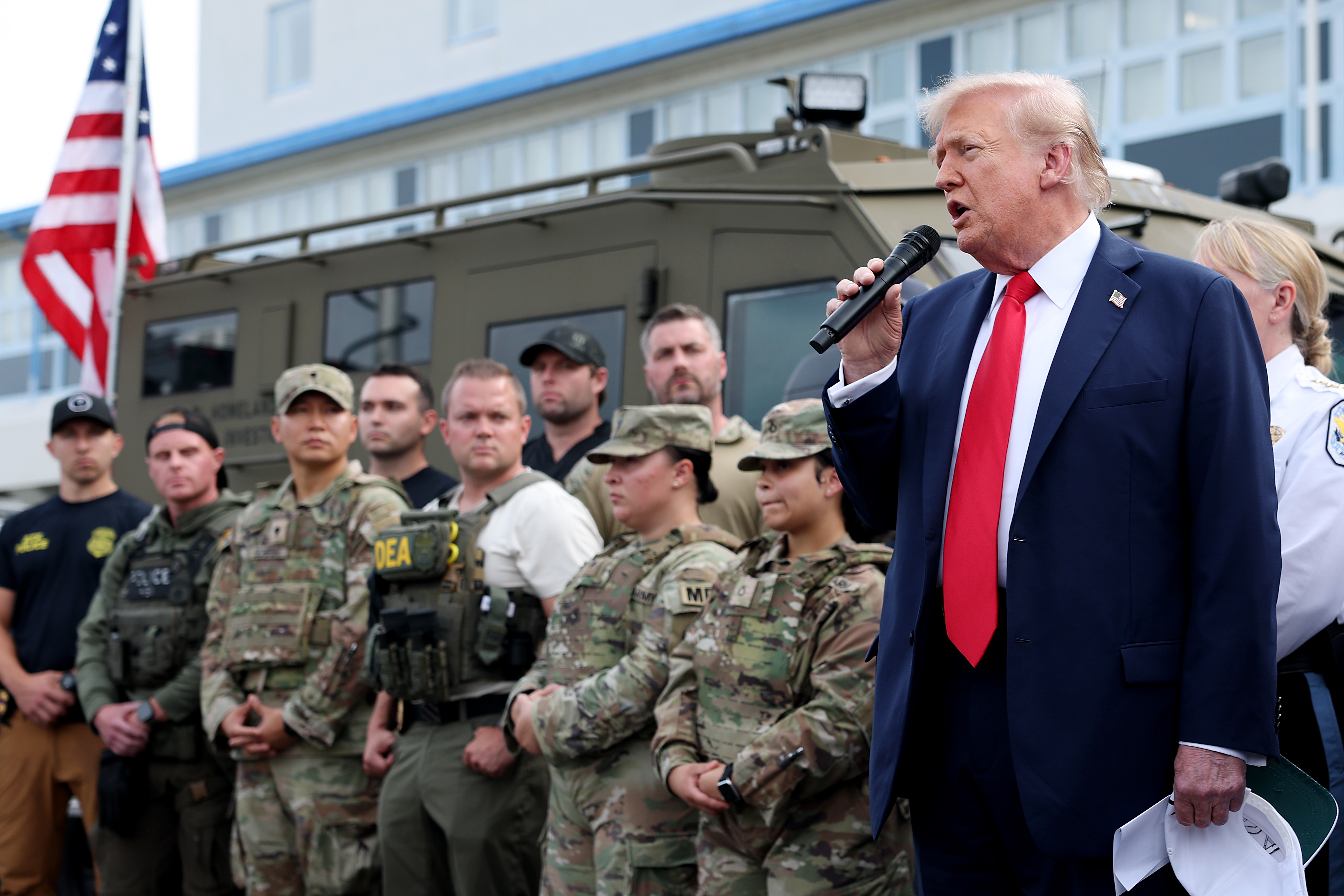 Public figure speaks into a microphone while standing in front of military personnel and law enforcement officers. American flag visible
