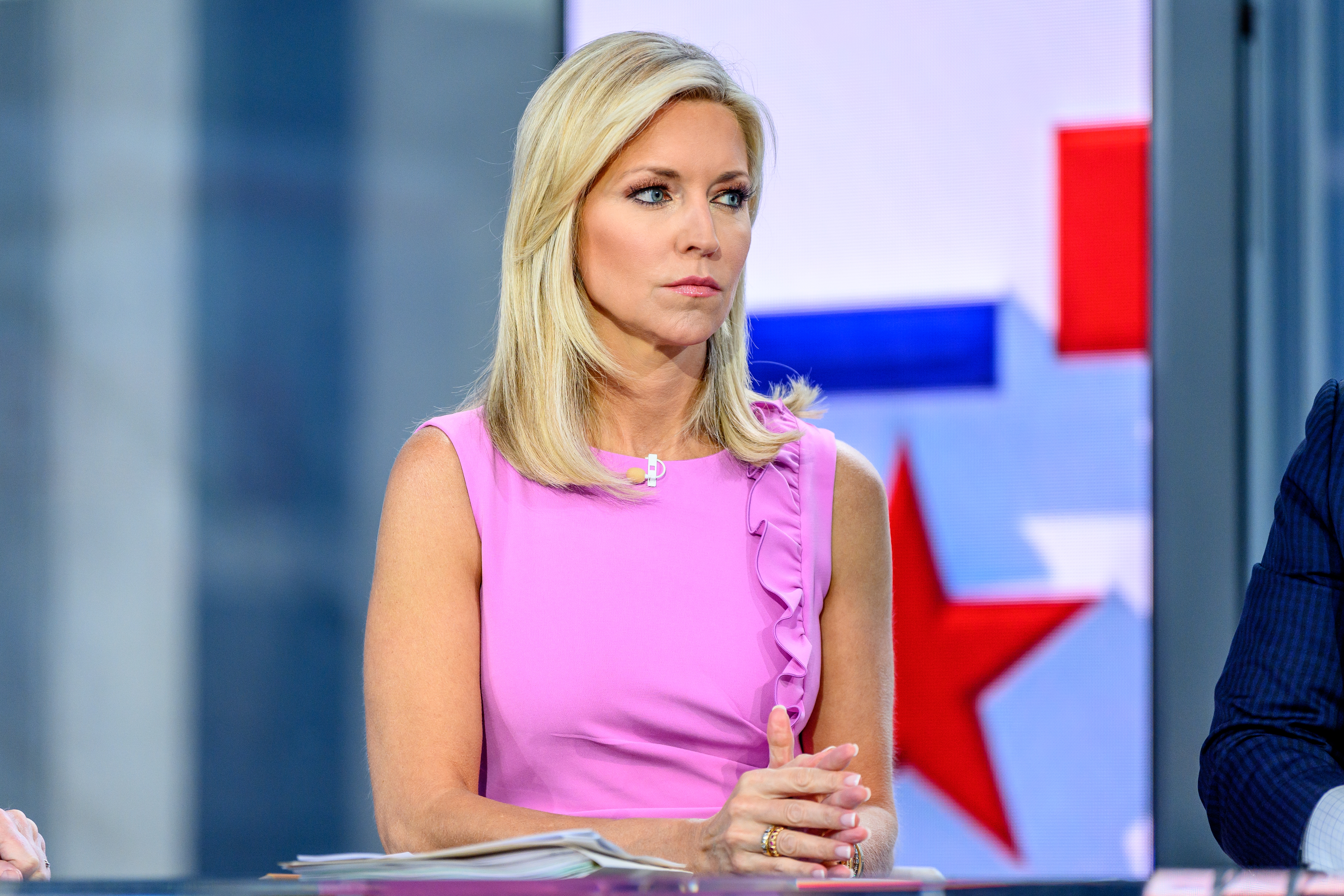 Person seated at a news desk, wearing a sleeveless ruffled dress, appears attentive while listening