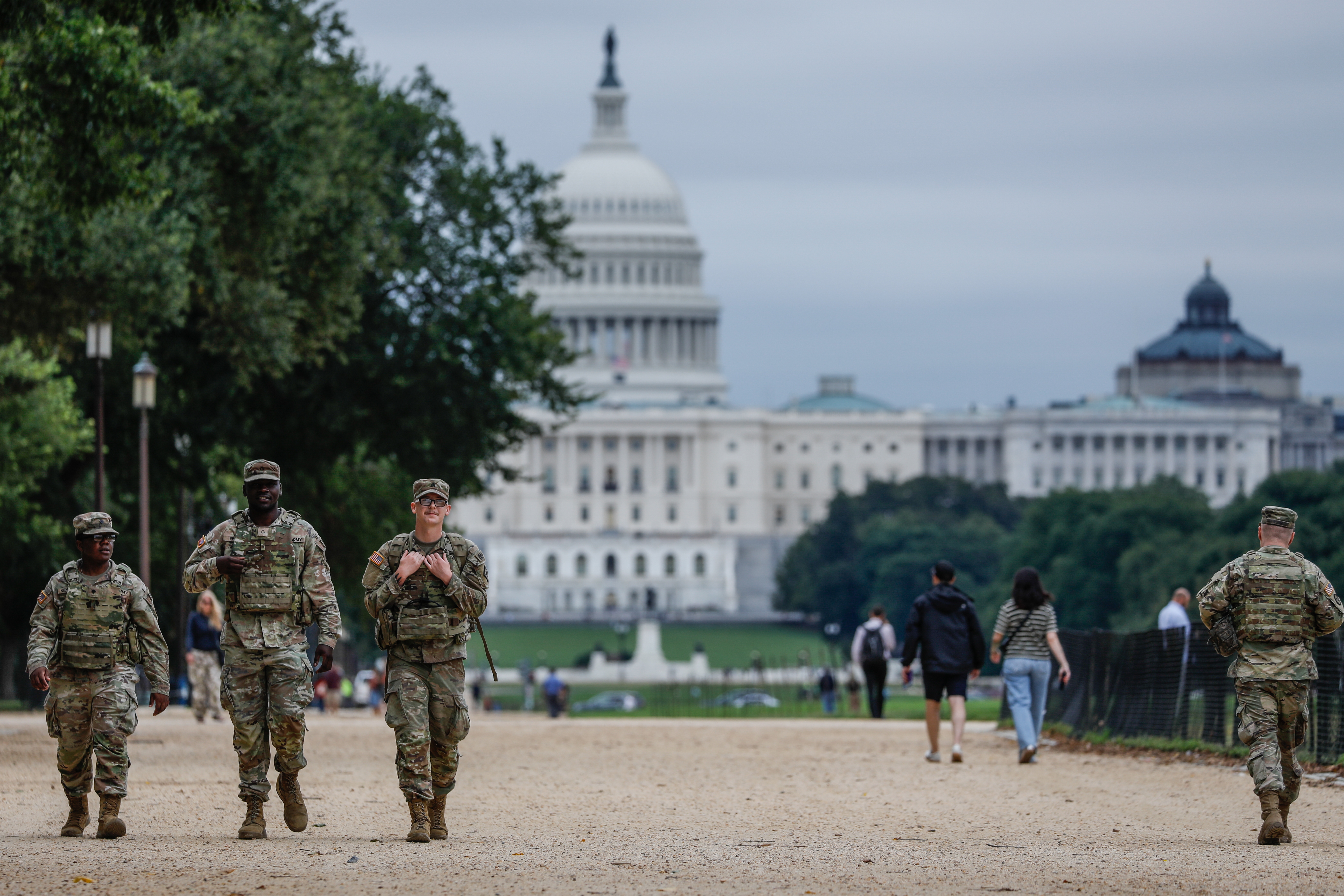 Five soldiers walking on a path in front of the U.S. Capitol building, surrounded by trees and pedestrians