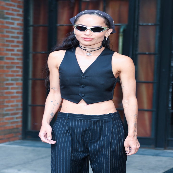 Person in pinstripe trousers, sleeveless vest, sunglasses, and bandana. Minimalist accessories and tattoos visible. Urban background