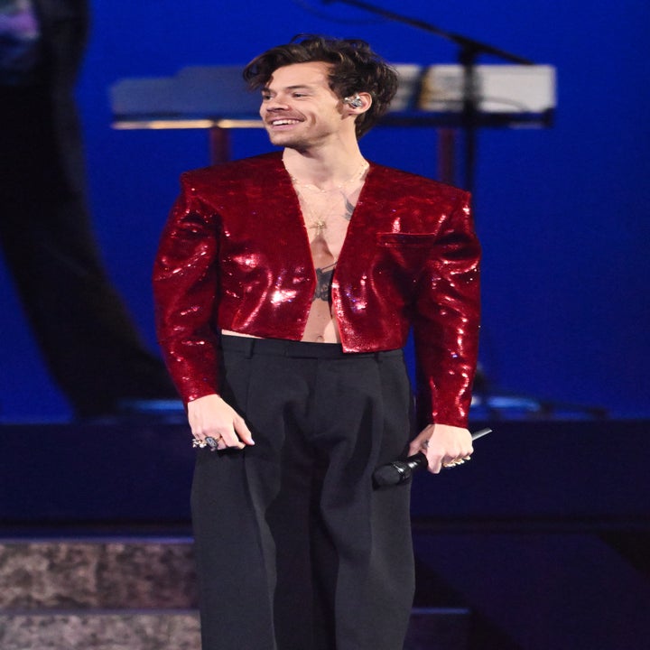 Person on stage wearing a shiny red cropped jacket and black high-waisted pants, holding a microphone, smiling