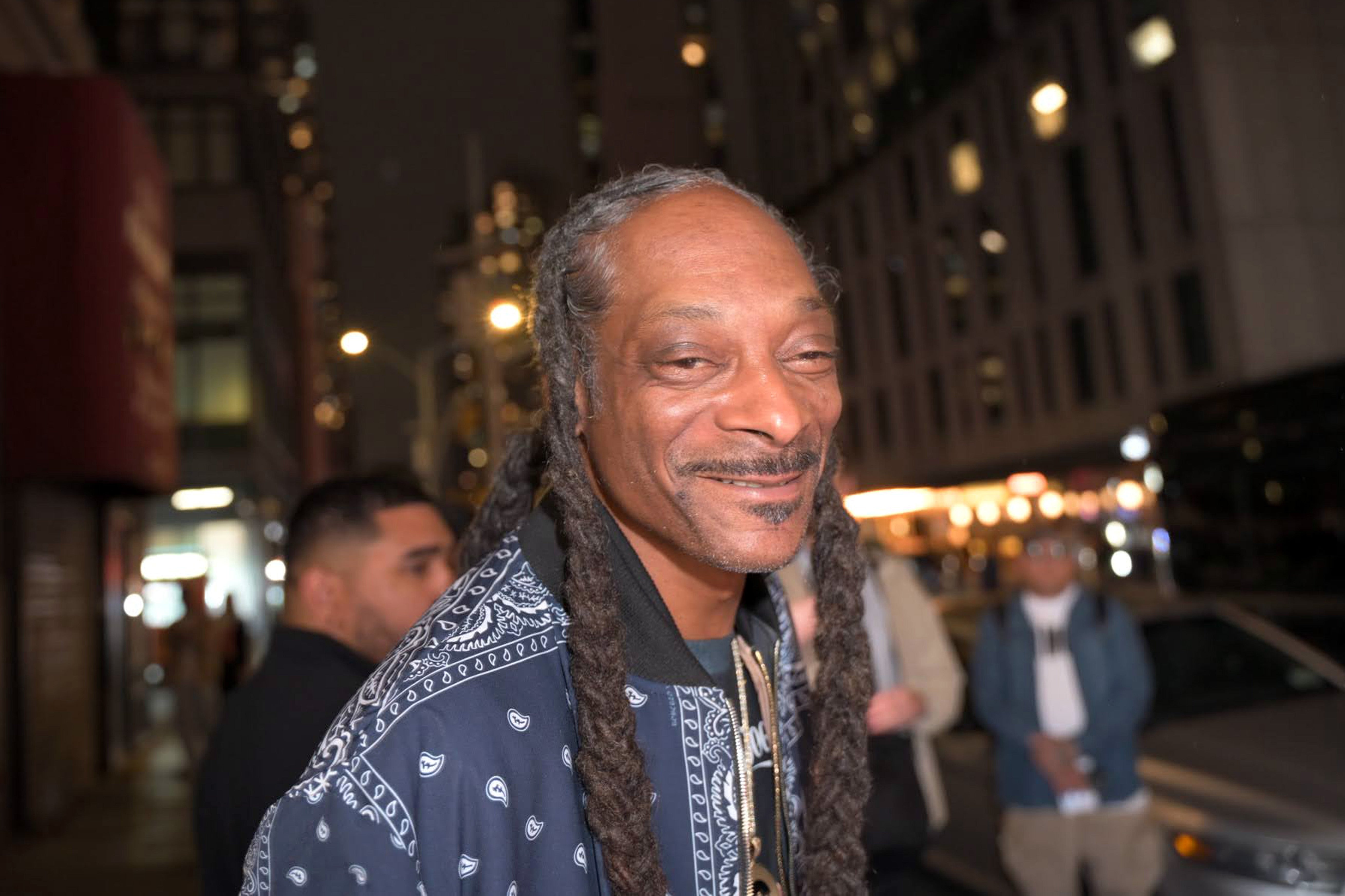 Snoop Dogg with long braids and a patterned jacket smiles on a city street at night