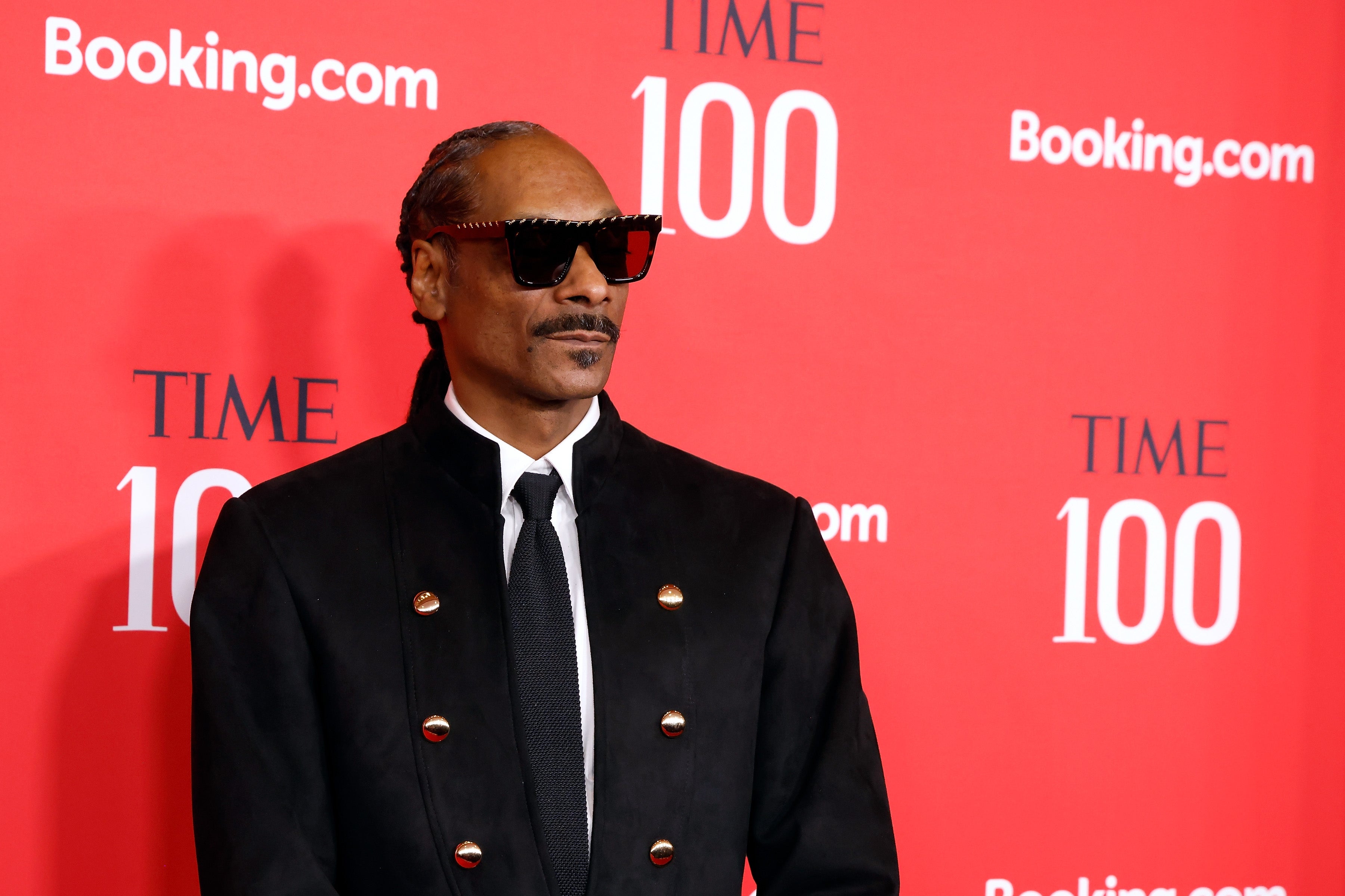 Snoop Dogg in a sleek black double-breasted suit with large buttons and sunglasses at a red carpet event with "Time 100" and "Booking.com" logos