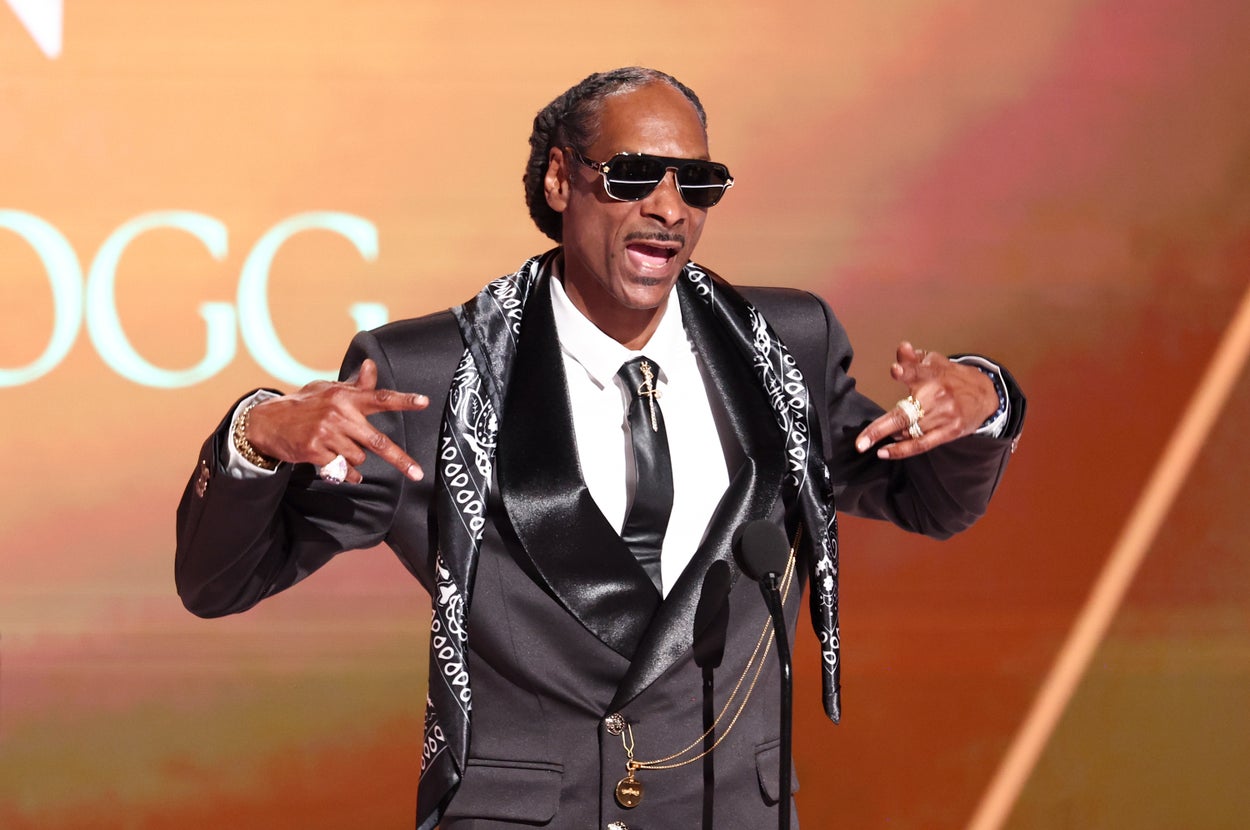 A man in a black suit with a patterned scarf gestures animatedly on stage, wearing sunglasses