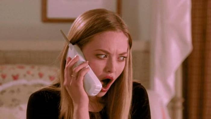 Person with long hair, eyes wide and mouth open, appearing shocked or surprised while holding a cordless phone to their ear indoors