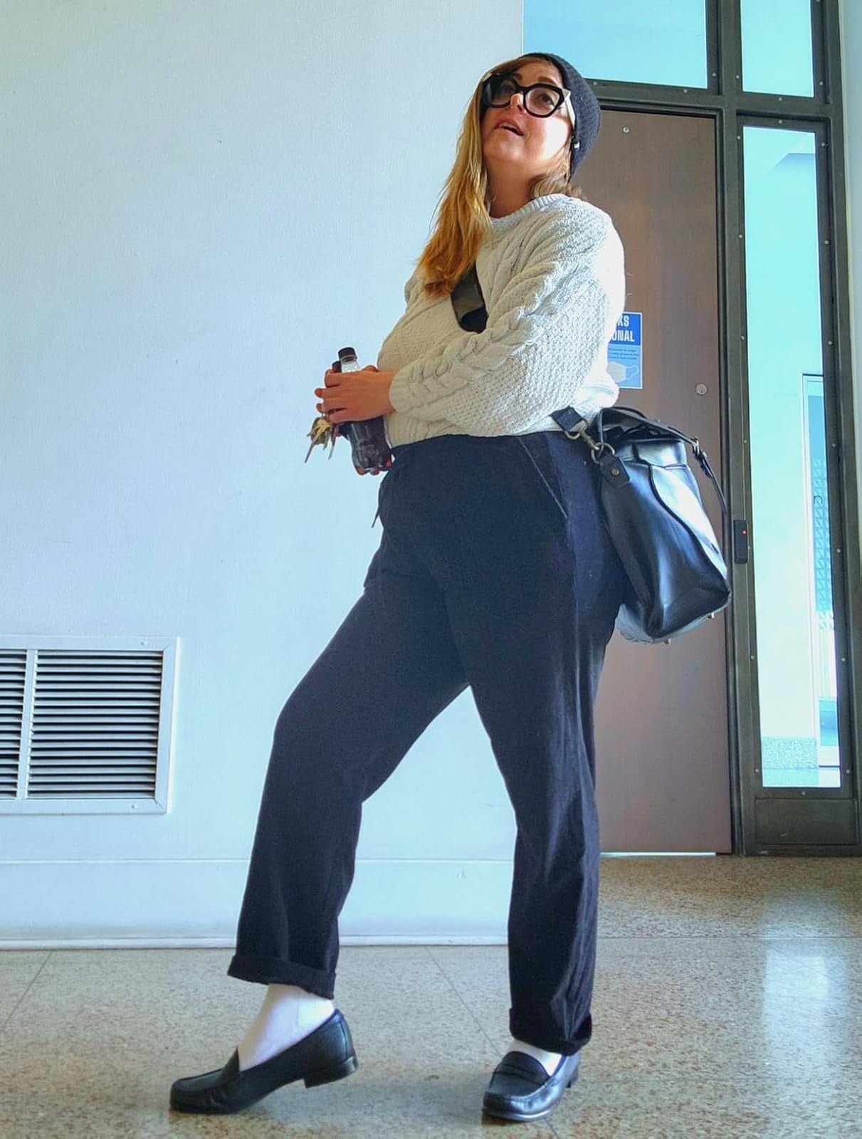 Person successful  casual achromatic  pants, sweater, hat, and loafers holding a portion  and keys. They person  a crossbody bag. Indoors against a neutral background