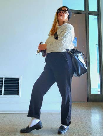Person successful casual achromatic pants, sweater, hat, and loafers holding a portion and keys. They person a crossbody bag. Indoors against a neutral background