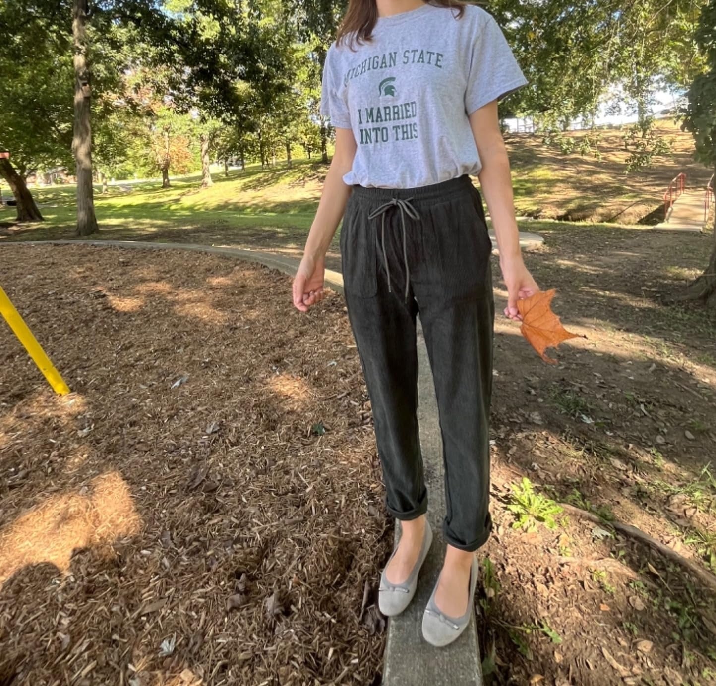 Person wearing a casual T-shirt and pants stands connected  a woody  borderline   outdoors holding a leaf