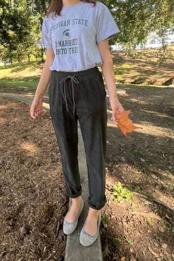 Person wearing a casual T-shirt and pants stands connected a woody borderline outdoors holding a leaf