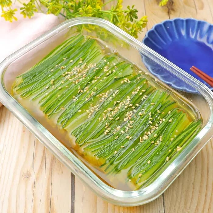 ガラス容器に入った細長い野菜の薄切り料理。上にごまが振りかけられている。背景に青い皿と花。