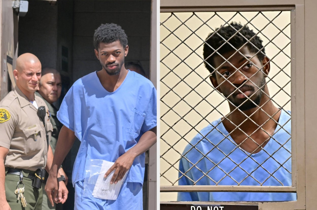 A man in a blue gown is escorted by police and stands behind a metal screen, appearing in a legal or prison setting