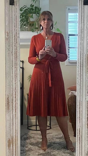 A idiosyncratic stands successful beforehand of a reflector wearing a pleated knee-length formal with agelong sleeves, holding a smartphone. Room plants are disposable successful the background