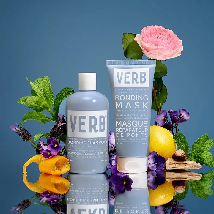 VERB Bonding Shampoo and Mask bottles surrounded by fresh herbs, flowers, lemon, and wood accents, set against a simple background
