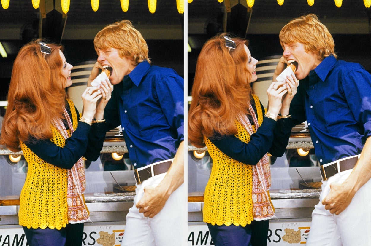 Two people happily share a sandwich at a food stall, both smiling and engaged. They're casually dressed, enjoying a lighthearted moment