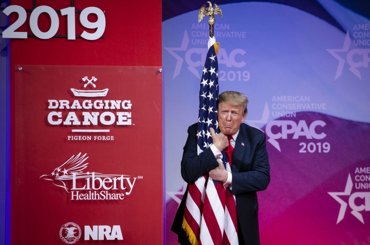 A person hugs an American flag on stage at a conference with logos including NRA and references to 2019 and Dragging Canoe, Pigeon Forge