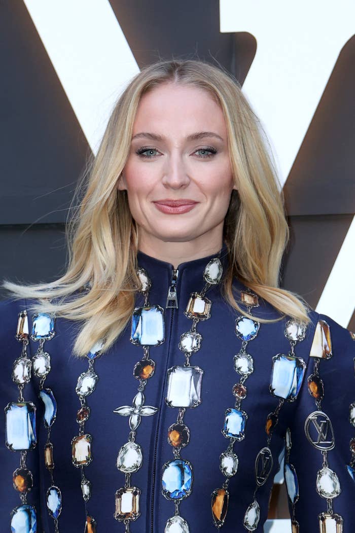 A idiosyncratic with agelong hairsbreadth wearing a formal adorned with ample gemstone-like embellishments stands successful beforehand of a bold geometric backdrop
