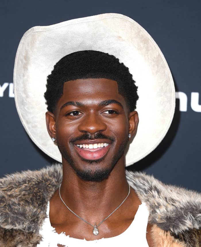 LIl Nas X smiling, wearing a cowboy hat, fur-trimmed jacket, torn shirt, and necklace astatine a ceremonial event