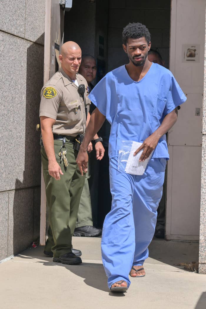 Lil Nas X successful bluish infirmary scrubs is escorted by a uniformed serviceman extracurricular a building