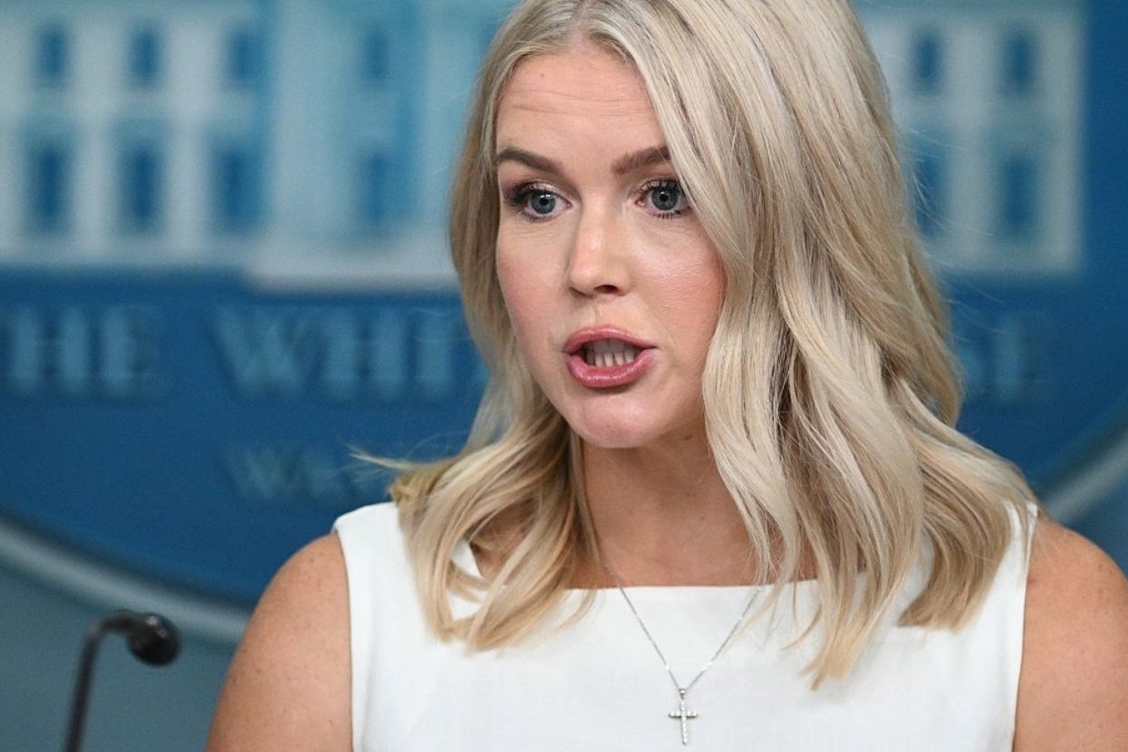 A woman with shoulder-length hair and a white sleeveless top speaks at a podium, with a blurred background of an official setting
