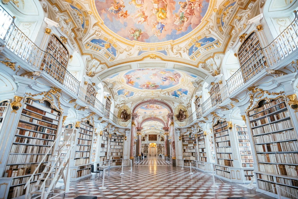 Ornate library interior with tall shelves, grand archways, and a detailed ceiling mural featuring artistic figures and architectural embellishments