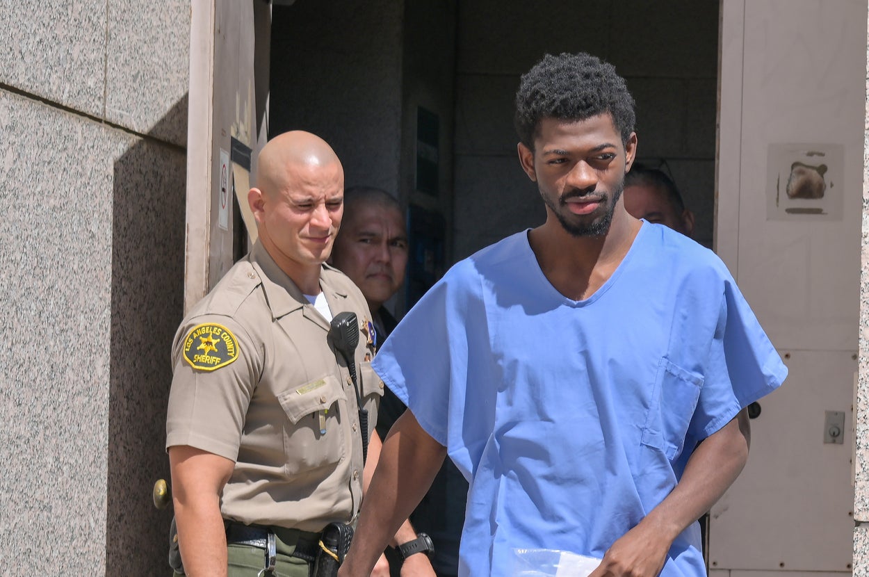 A man in blue scrubs and sandals leaves a building escorted by a law enforcement officer