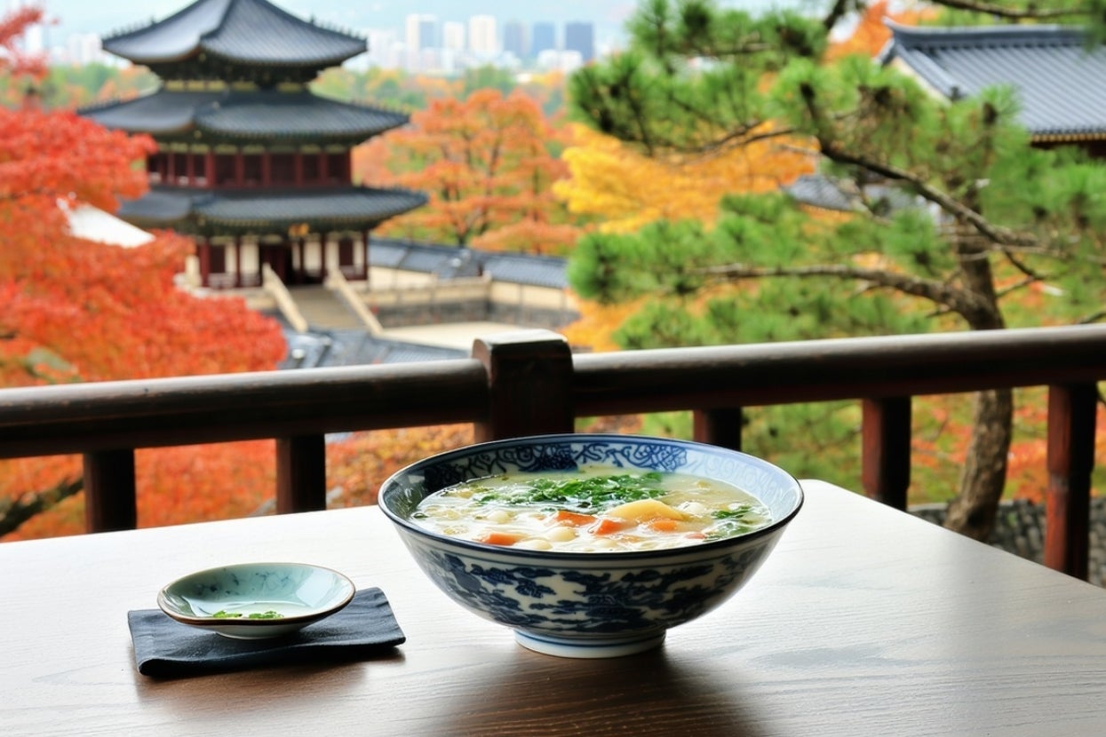 A bowl of soup with vegetables on a wooden table overlooks a scenic view of a traditional pagoda and colorful autumn trees