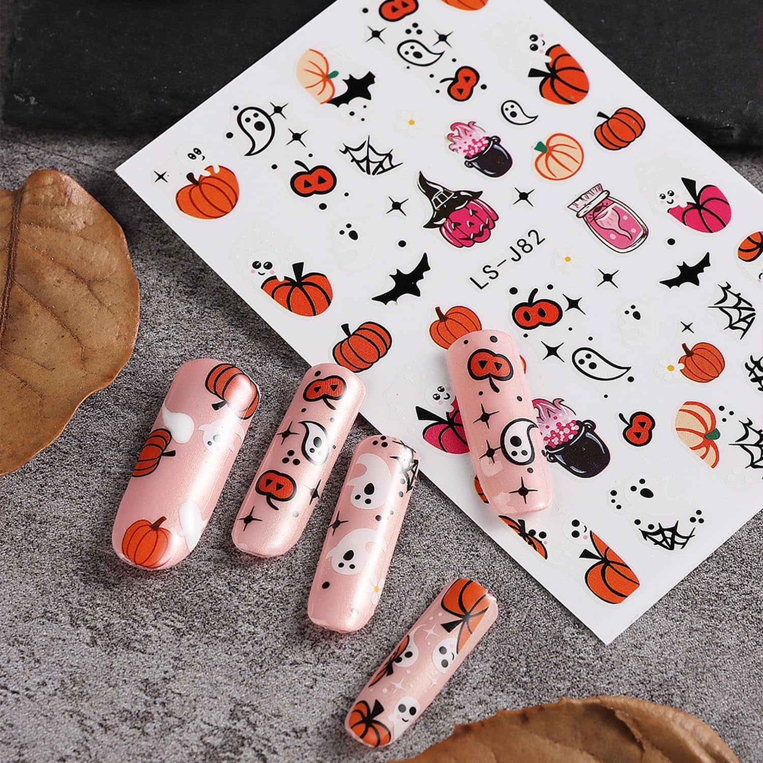 Five artificial nails adorned with Halloween-themed designs featuring ghosts, pumpkins, and bats next to a sticker sheet with similar motifs.