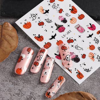 Five artificial nails adorned with Halloween-themed designs featuring ghosts, pumpkins, and bats next to a sticker sheet with similar motifs.