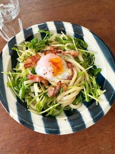 皿に盛り付けられたウドン、温泉卵、ベーコン、豆苗が添えられた料理。