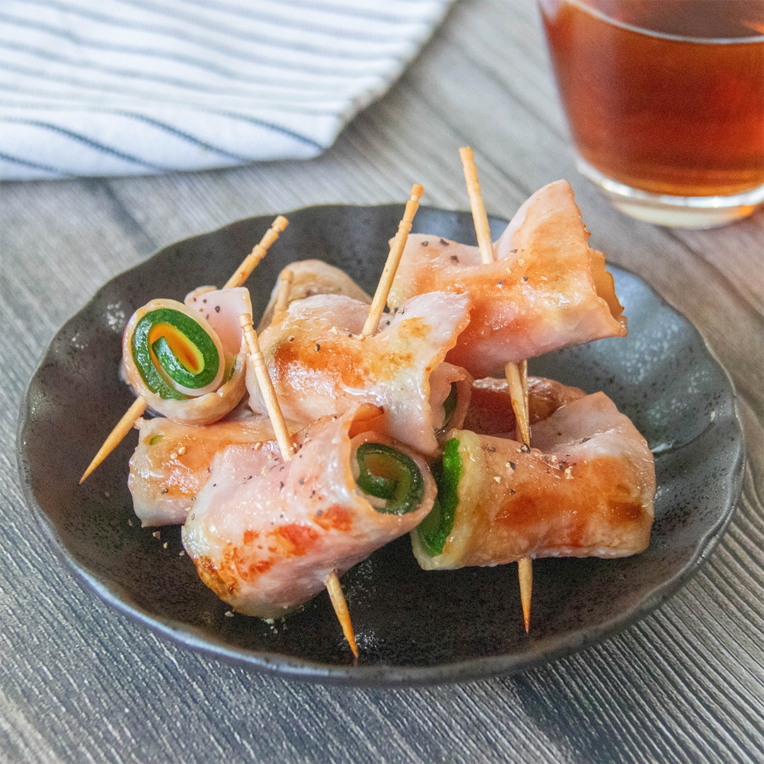 料理の写真。ベーコンで巻かれたズッキーニの串刺しが黒い皿に盛られている。黒コショウがかかっており、バックには飲み物が見える。