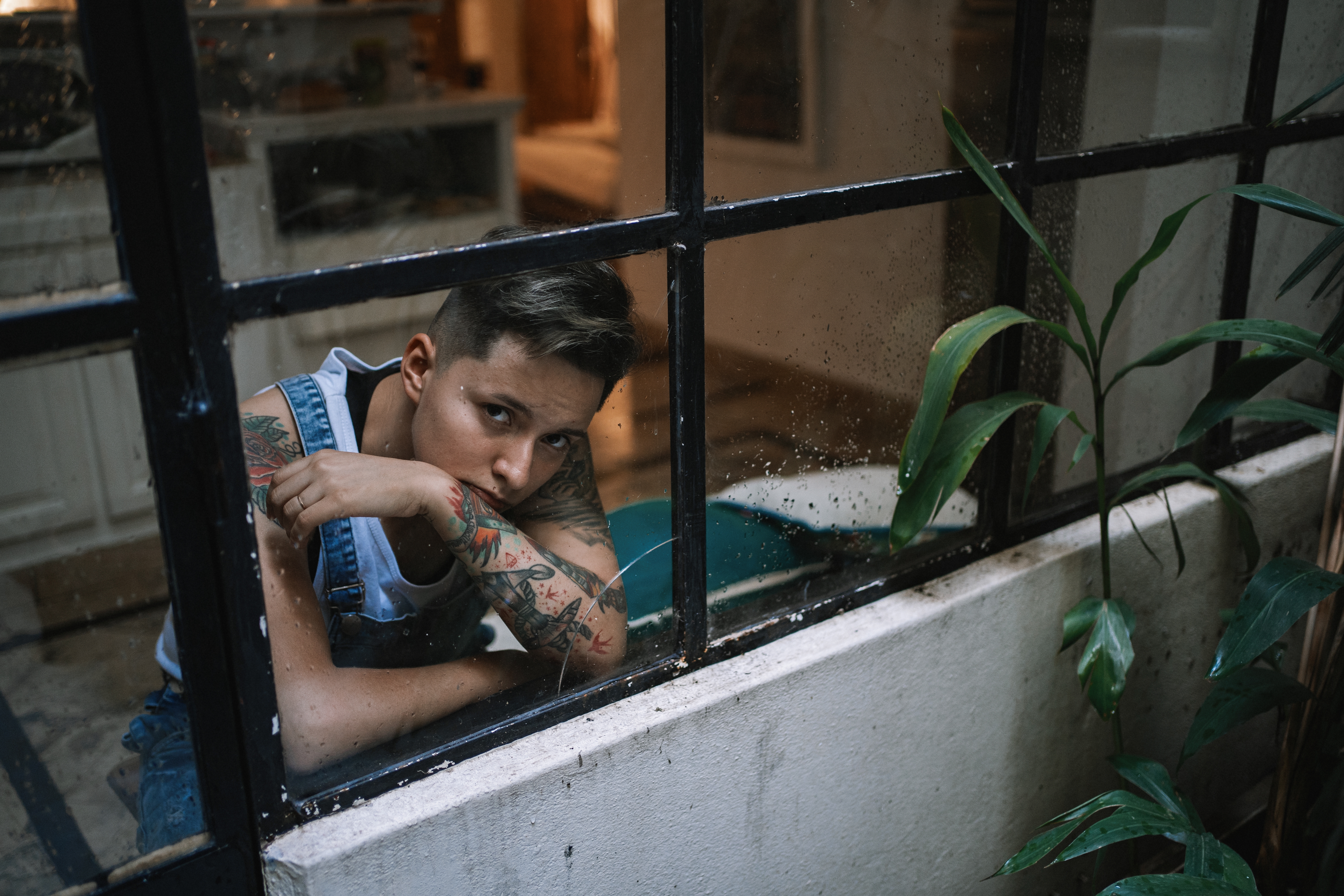 Person med ermet tatoveringer stirrer pensivt gjennom et vindu, iført denim -kjeledress, omgitt av innendørs planter i koselig setting