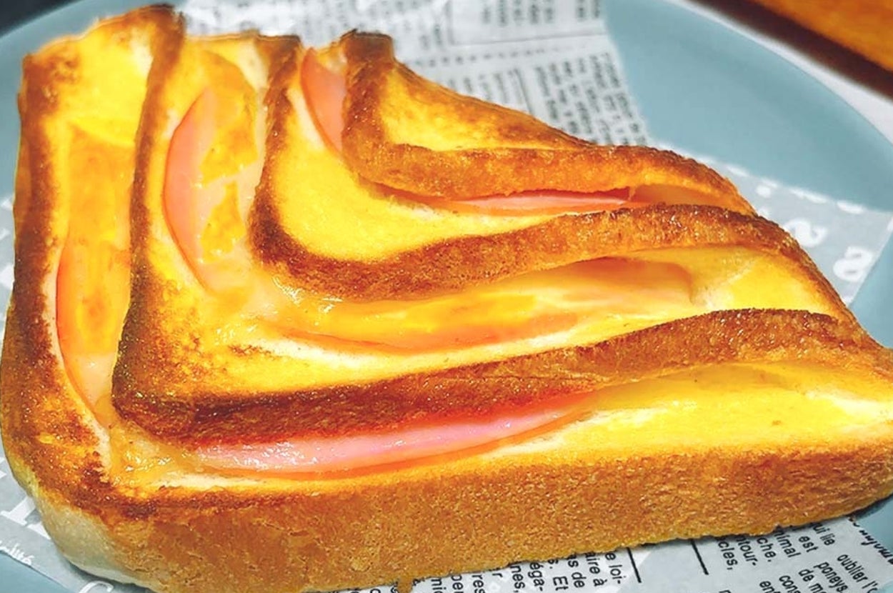 トーストにハムとチーズが挟まれたサンドイッチが、新聞紙に似た紙の上に乗り、青い皿に盛られている。
