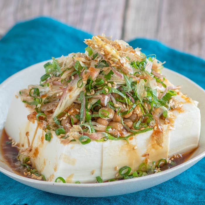 冷奴に刻みねぎと鰹節、ゴマが乗った料理。味付けに醤油がかかっている。