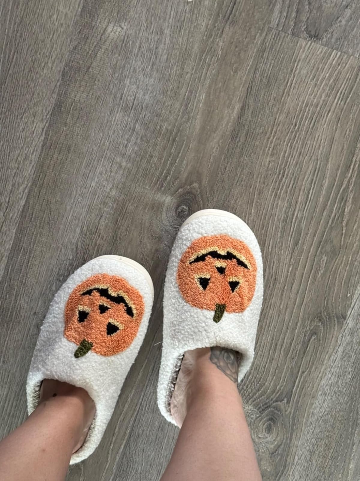 Feet wearing cozy slippers with a pumpkin face design, standing on a wooden floor.